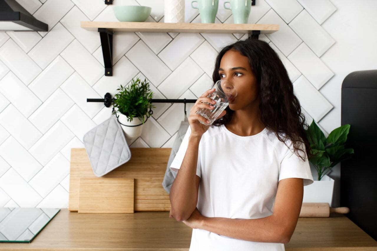 mulher bebendo água na cozinha para ilustrar texto sobre benefícios da água para a pele