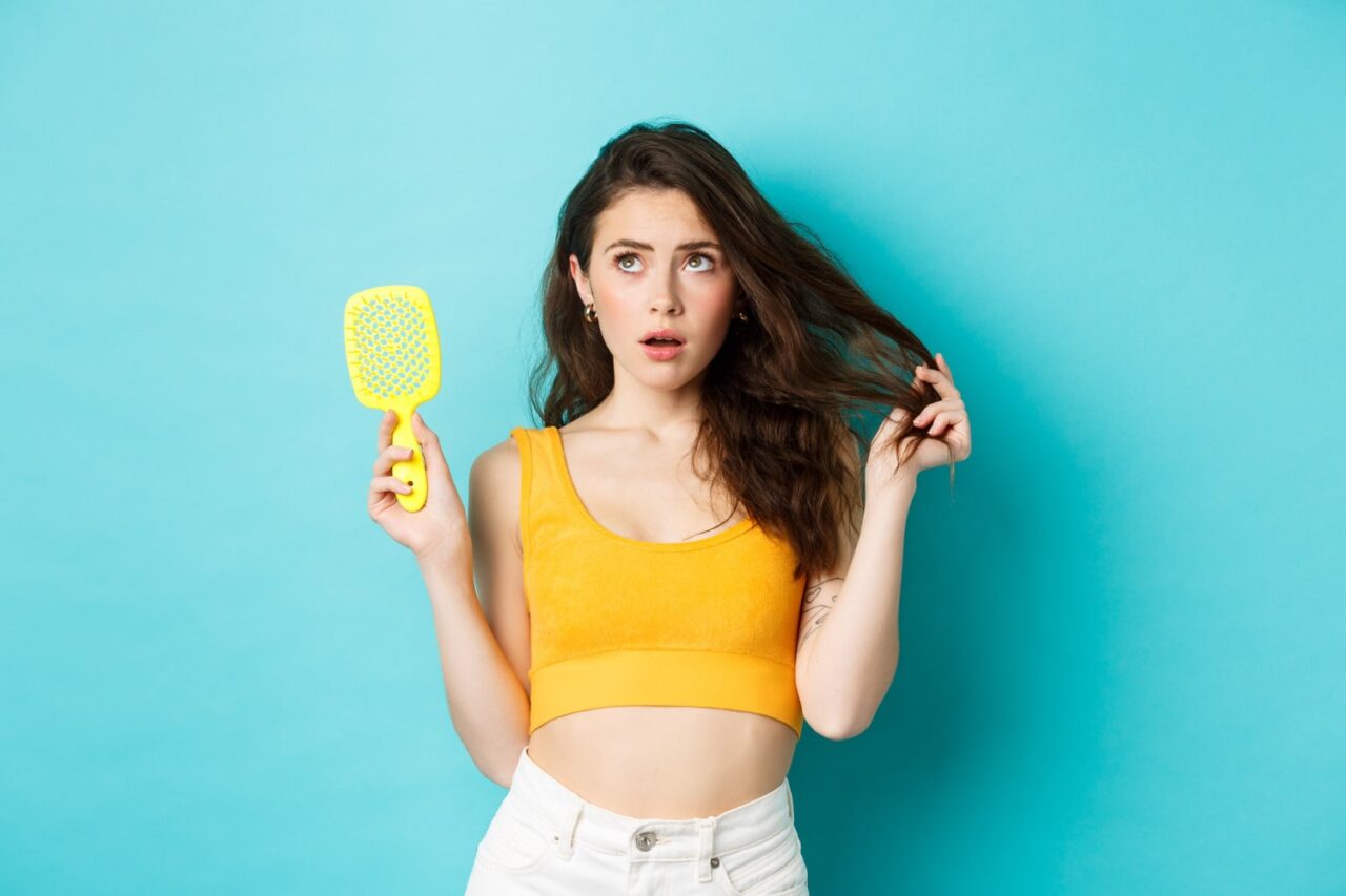 mulher com uma escova na mão e outra no cabelo com expressão de espanto para ilustrar texto sobre o que deixa o cabelo oleoso