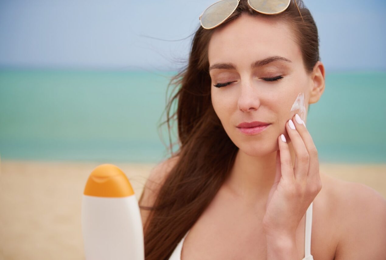 mulher na praia aplicando protetor solar no rosto para ilustrar texto sobre como saber qual fator de protetor solar usar
